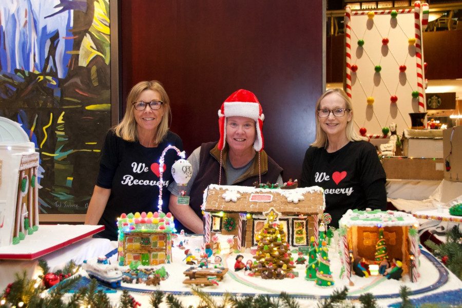 Three women, one in a Santa hat, two wearing t-shirts saying "We Heart Recovery" standing behind several ginger bread houses, picnic tables, Christmas trees, people and a ferry on railroad tracks.