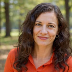 Smiling woman with wavy brown hair wearing a red shirt. Blurry trees are in the background. Nicole Fetterly - Orchard Dietitian