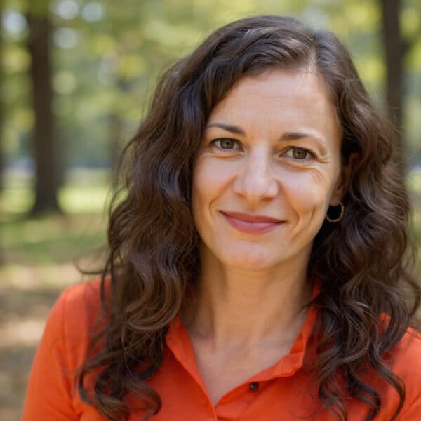 Smiling woman with wavy brown hair wearing a red shirt. Blurry trees are in the background. Nicole Fetterly - Orchard Dietitian